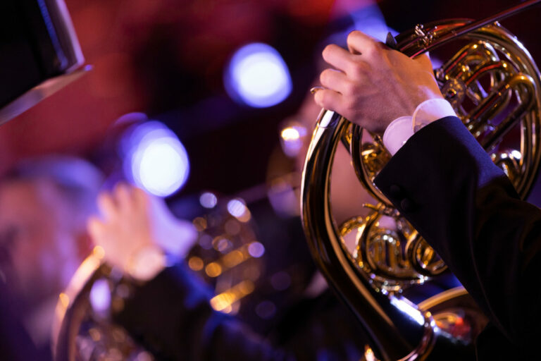 A French horn player playing his instrument in a classical symphony orchestra with another player in the background with the room flooded with colorful stage lights
